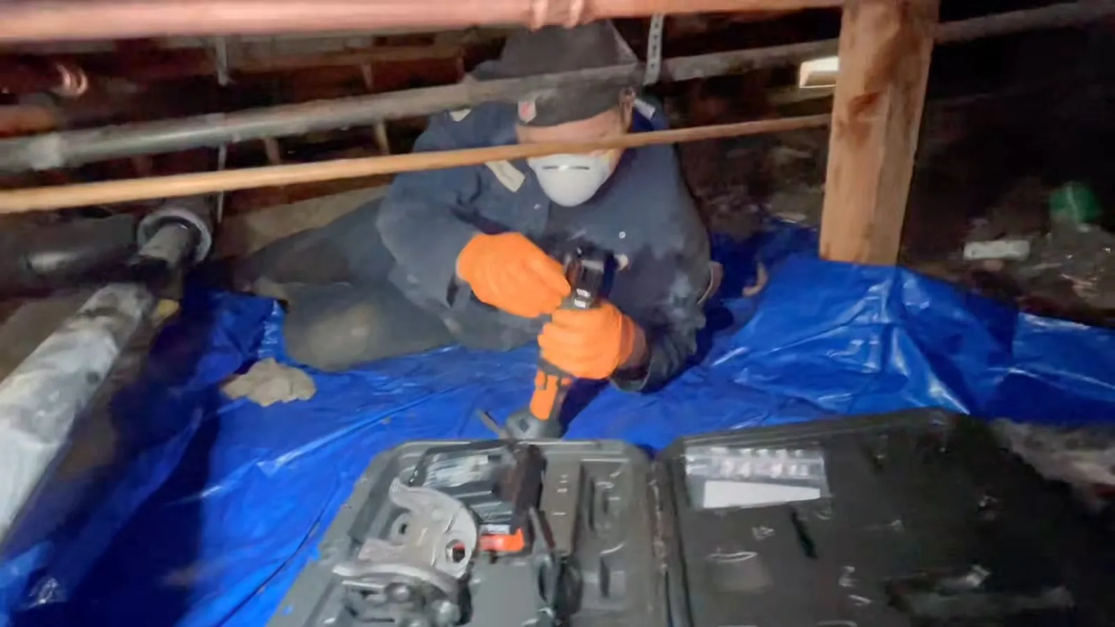 Worker in a mask and orange gloves kneeling on a blue tarp in a crawl space, tightening a fitting above an open tool case