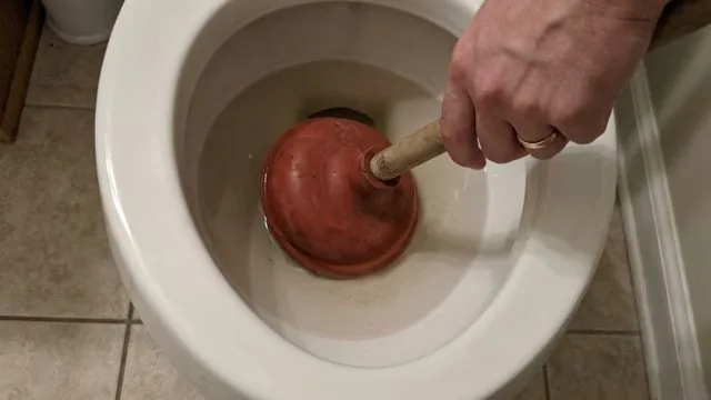 Hand pressing a red plunger into a toilet bowl on a tiled floor