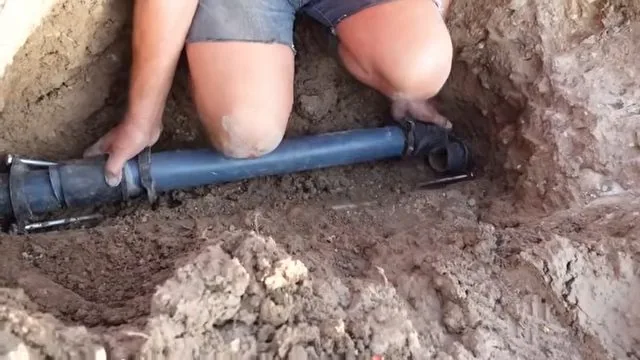 Person kneeling in a dug trench with both hands on a blue pipe, sliding a black coupling over a cut in the line