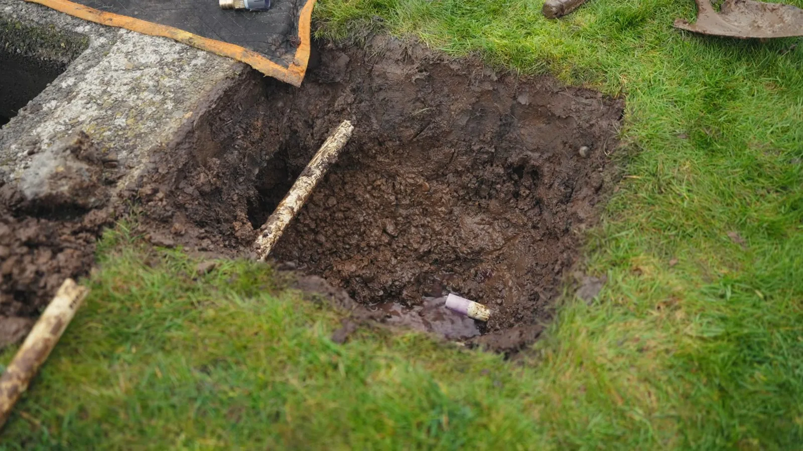 Small square pit dug in a lawn beside a concrete curb, with a wooden stick and a short pipe visible in the soil