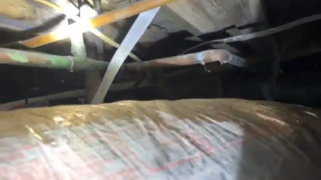 Camera view in a dark crawl space showing copper pipes, a metal strap and stained foil-faced insulation sagging below the floor