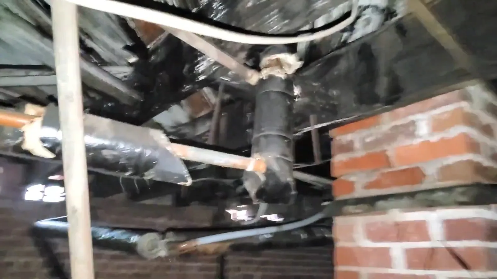 View under a floor of a pipe joint with rust staining, black sheeting and a brick pier beside it