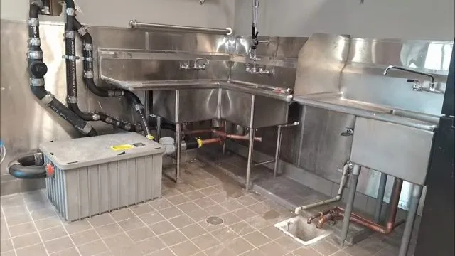 Commercial kitchen with stainless sinks, black drain pipes, a gray grease interceptor box on the floor and a floor drain in the tile