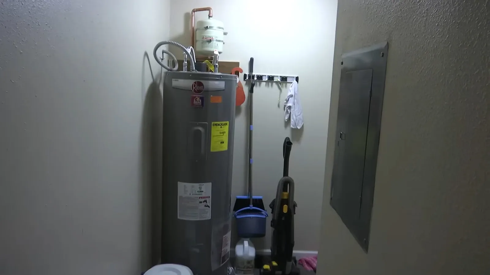 Gray tank water heater standing in a narrow utility room beside a wall-mounted electrical panel, with a mop and cleaning tools hanging nearby