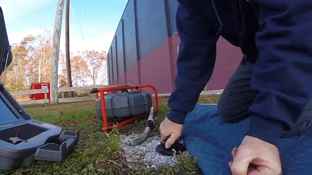 Person kneeling on grass feeding a cable into a ground cleanout, with a camera reel and monitor case beside him