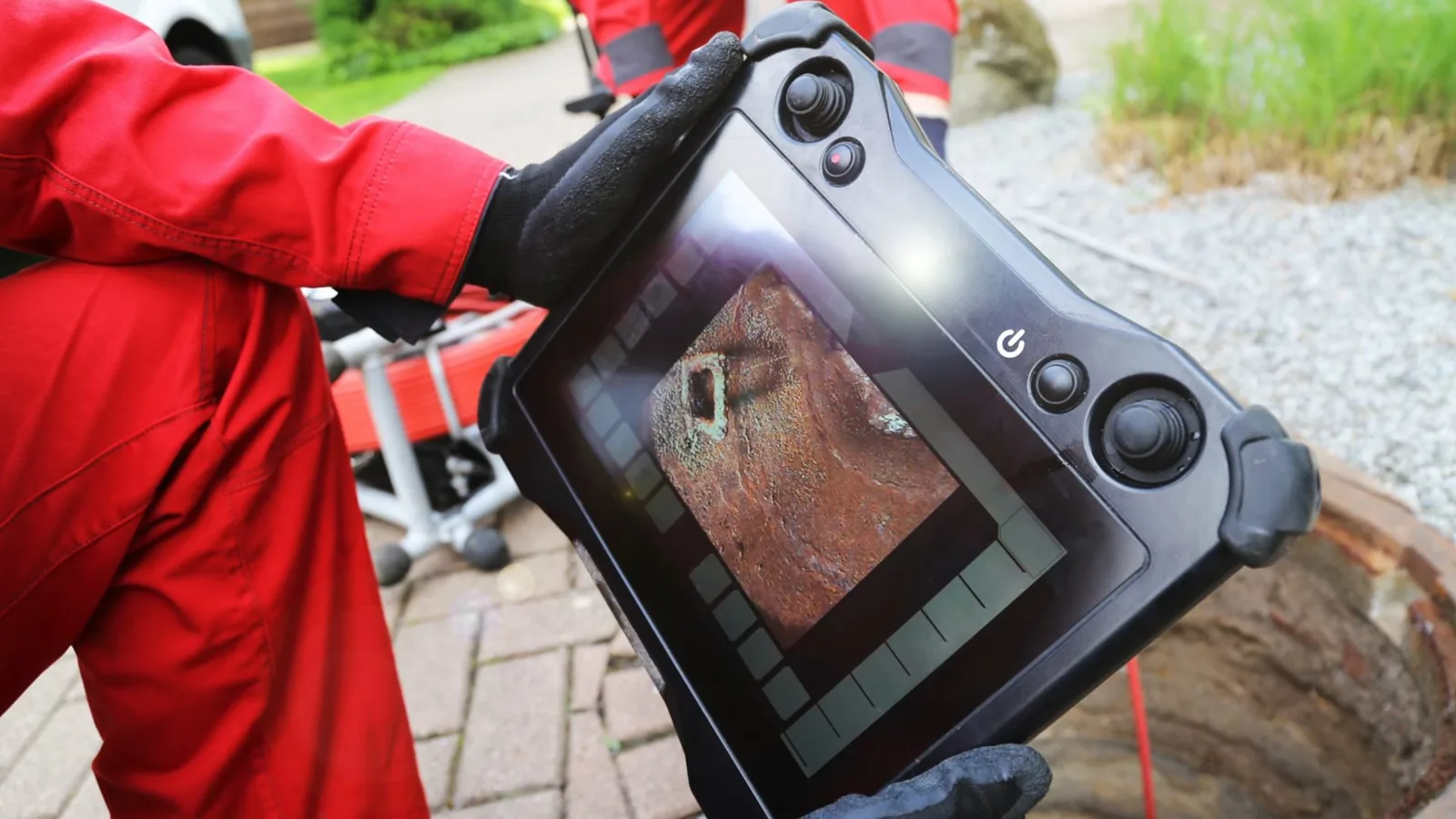 Technician in red coveralls holding a handheld sewer camera monitor over a paved area, with a cable reel behind