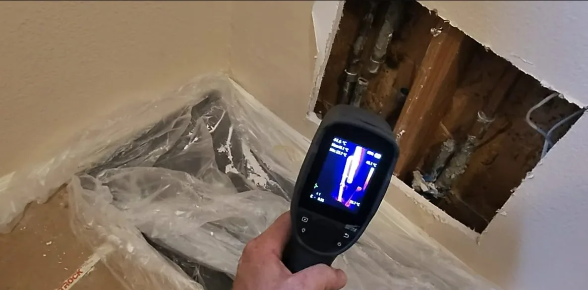 Hand-held thermal imaging camera pointed toward a wall opened to show pipes, with plastic sheeting on the floor below