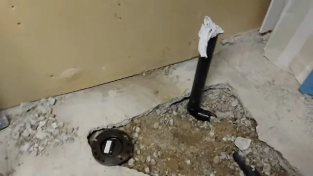 Black drain pipe and a toilet flange set in a broken-up concrete floor, with a vent pipe rising beside it and rubble around