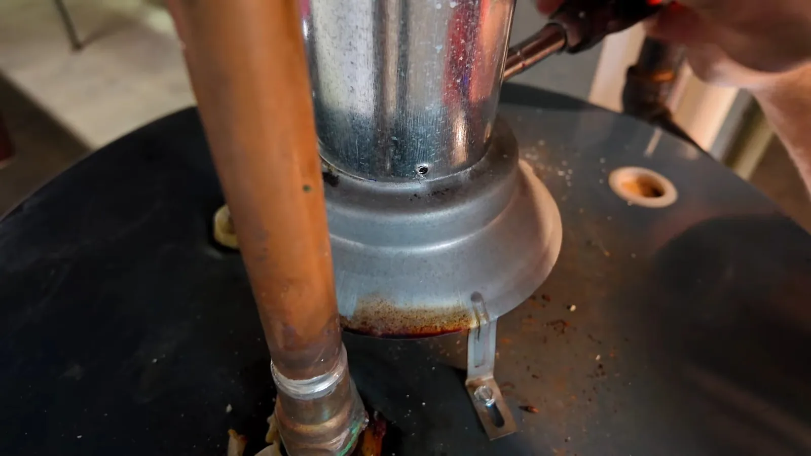Close view of the top of a water heater with a galvanized vent pipe and a copper line rising from it, and a hand holding a tool at upper right