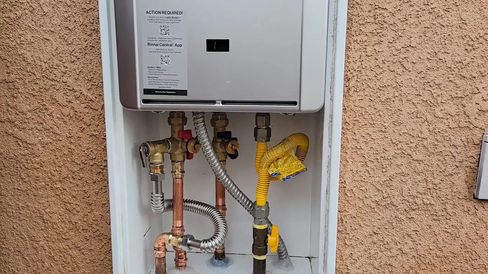 Wall-mounted tankless water heater on a stucco wall with copper pipes, shutoff valves, a braided hose and a yellow gas line below it