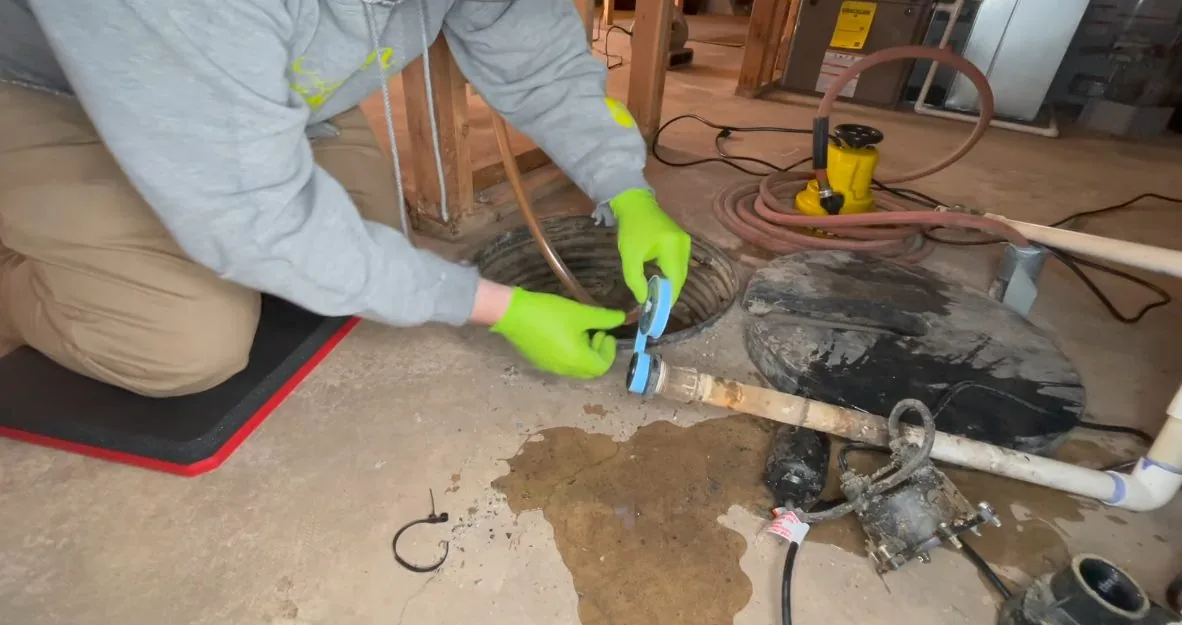 Worker in green gloves kneeling on a damp concrete basement floor beside a sump basin and its discharge pipe